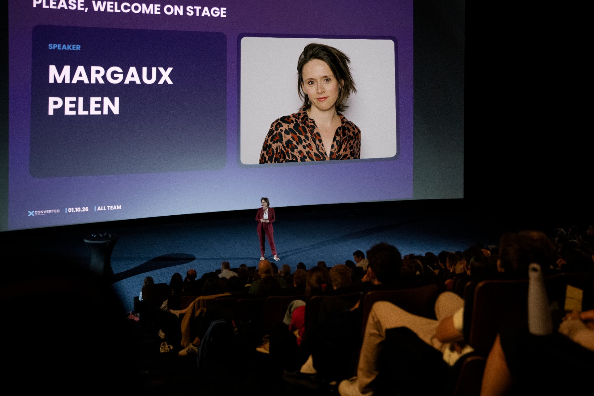 Margaux Pelen on stage at a Converteo all-team event, a screen behind her reading Please, Welcome on Stage